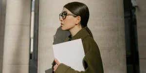 Женщина в зелёном пальто стоит у бетонной колонны