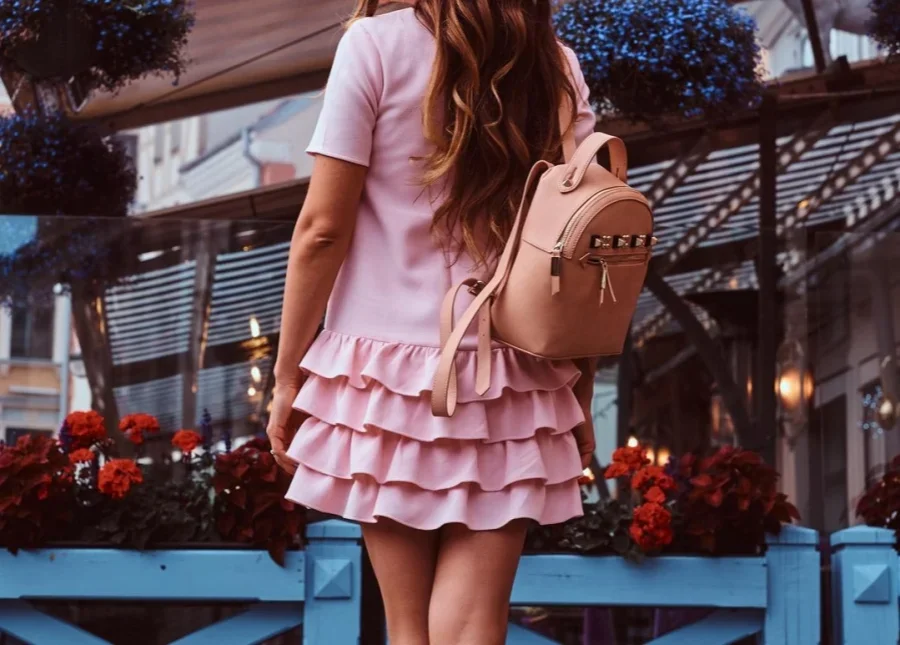 A brown-haired woman in a pink dress and a backpack stands near a terrace cafe, her back view