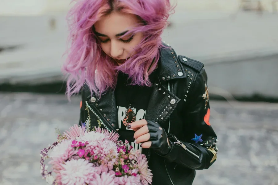Eine stylische junge Frau mit lila Haaren hält einen Blumenstrauß in der Hand und trägt eine schwarze Lederjacke im Freien