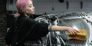 A young woman cleans a black car with a sponge and soap in a garage setting, showing care and attention