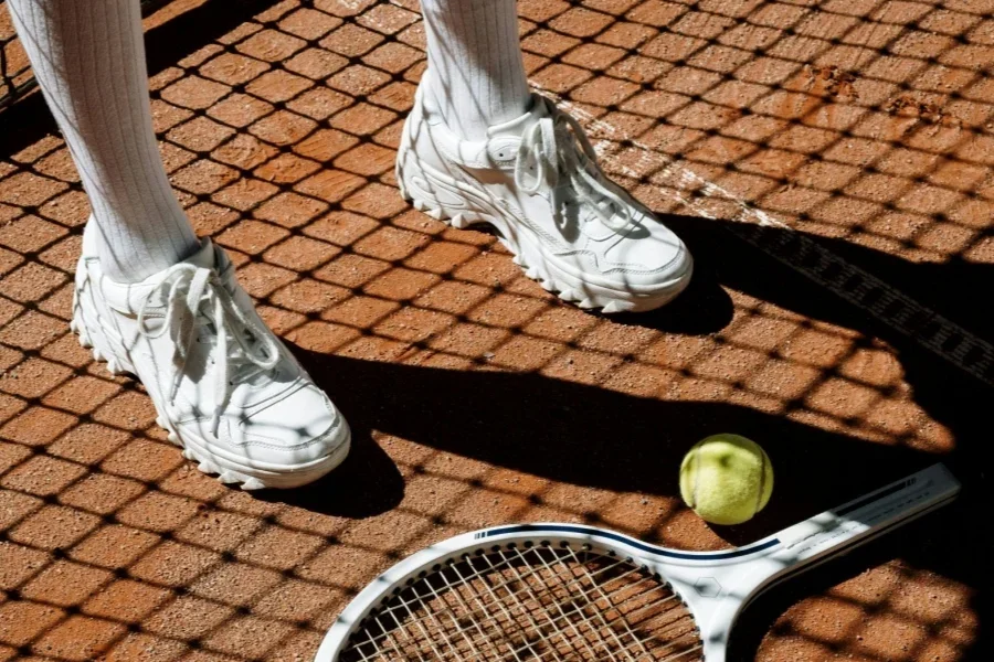Person trägt weiße Turnschuhe und steht neben einem Tennisschläger