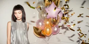 Stylish woman in silver dress celebrating birthday with balloons indoors