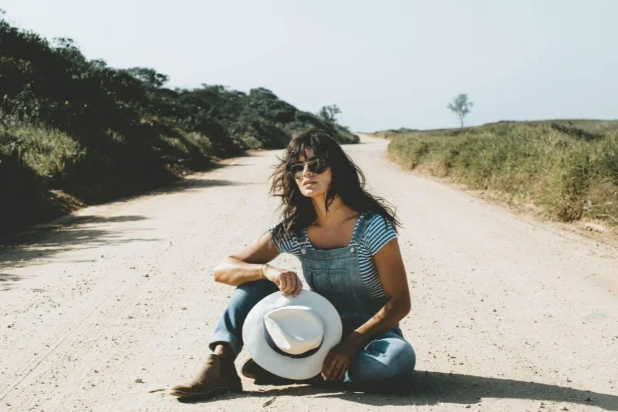 Woman Wearing Denim Overalls and Sunglasses Sitting on the Road in the Countryside