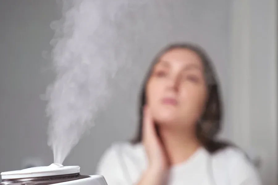 Una joven morena aplica crema humectante sobre la piel seca de su rostro sentada en una habitación cerca de un humidificador que vaporiza el aire en primer plano.