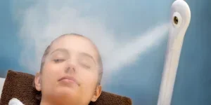 up portrait of young woman with eyes closed having thermal steam treatment on face. Girl wearing white gown laying on couch in spa