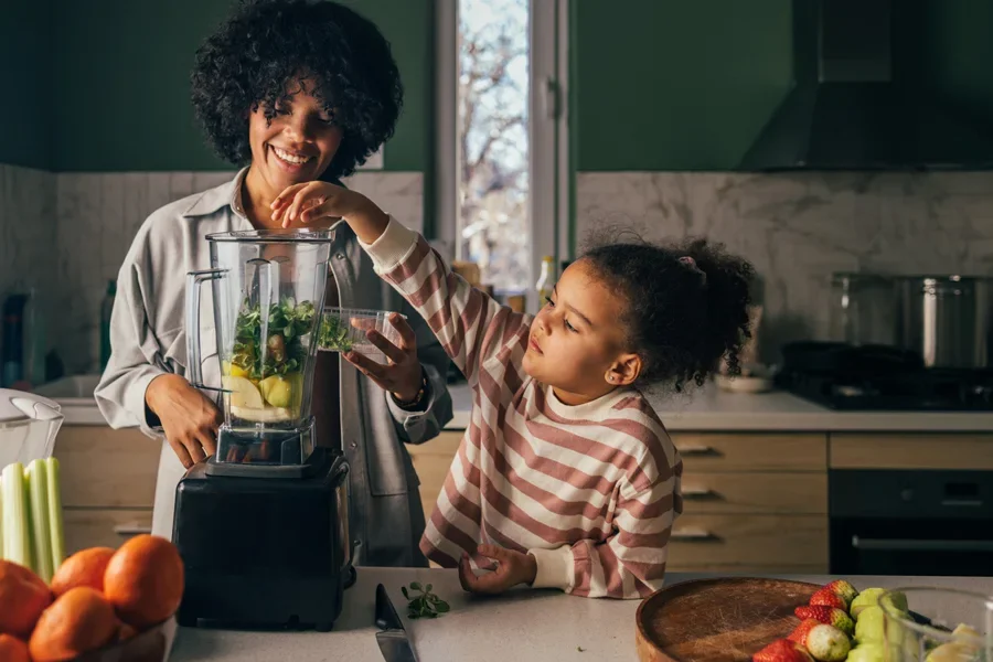 Улыбающиеся мать и дочь вместе готовят полезный смузи